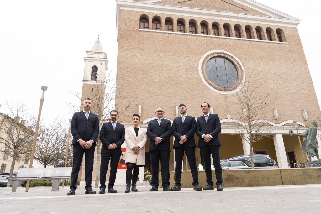 Cinque uomini in abito scuro e una donna in cappotto chiaro davanti a una chiesa in mattoni con rosone e campanile
