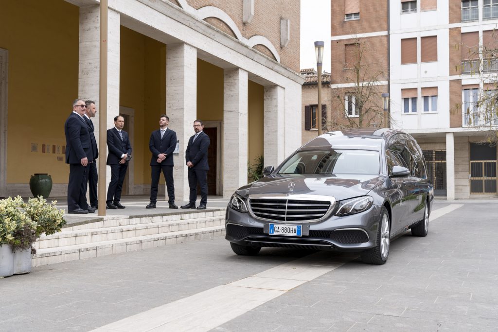 Cinque uomini in abito scuro sotto un portico con colonne in travertino accanto a un carro funebre Mercedes grigio