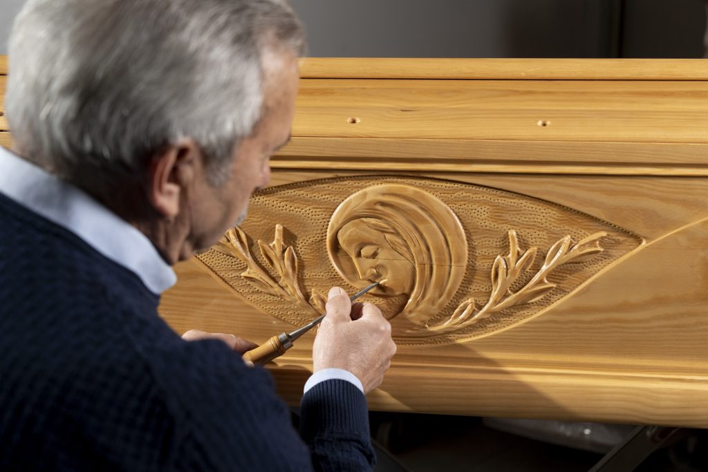 Artigiano dai capelli grigi intaglia con sgorbia un volto di Madonna tra rami d'ulivo su una bara in legno chiaro